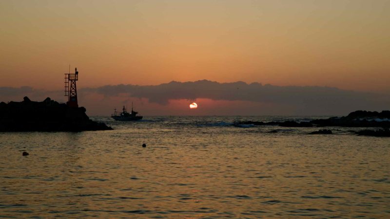 A fishing boat departs Muam Port at sunrise, silhouetted against a soft, glowing sky.