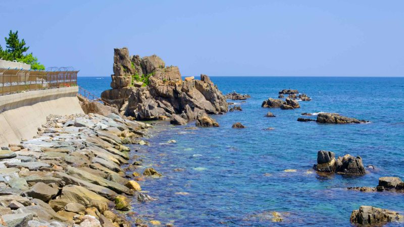 A coastal rock formation juts out just off the shore near Geojin Port, where a roadway edged by barbed wire fencing snakes along the coastline.