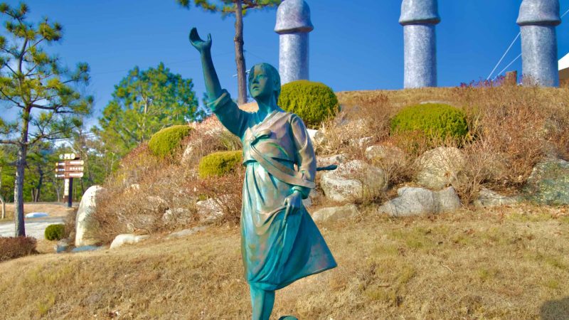 A stone sculpture of the young lost maiden, in the same iconic pose, set against a hillside and surrounded by penis zodiac statues in Haesindang Park.