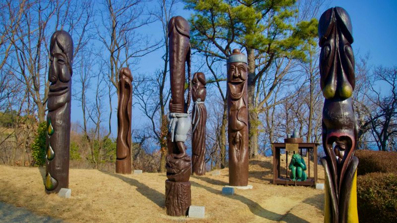 A wide-angle shot capturing an array of tall wooden penis sculptures with faces, and a depiction of a woman in a cage, chained in between, at Haesindang Park.