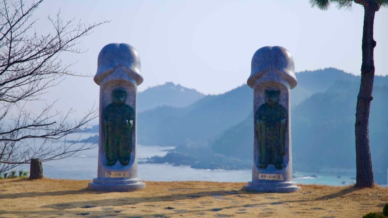 A close-up of two stone penis sculptures at Haesindang Park, with the serene East Sea and mountainous landscape forming a picturesque backdrop.