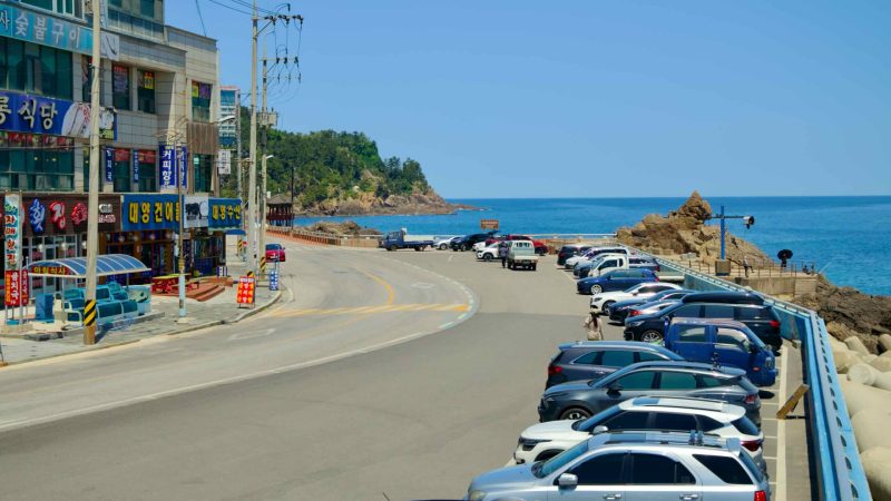 A view towards Lion Rock from Isabu Plaza, showcasing the coastal road lined with parked cars and local shops.