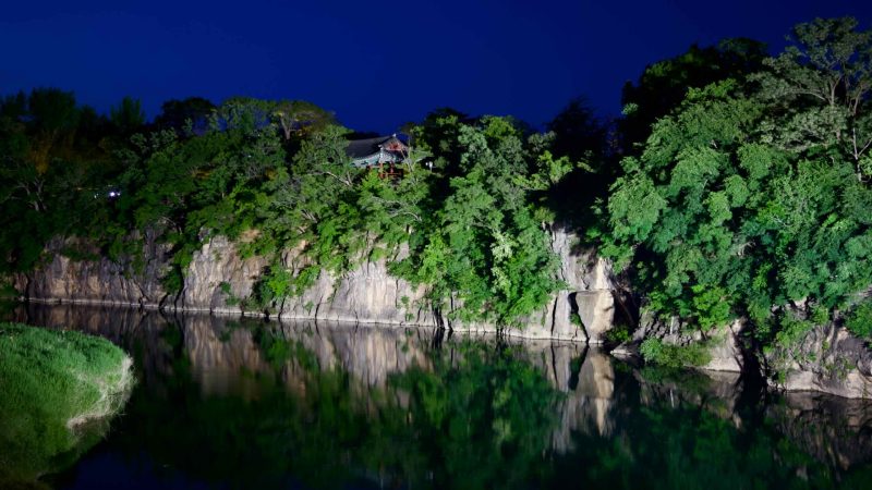 Jukseoru Pavilion illuminated at night, perched on a rocky clifftop, reflecting in the calm waters of the Osip Stream.