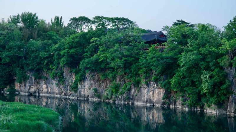 A view of Jukseoru Pavilion perched on a rocky clifftop, surrounded by lush green foliage, overlooking the calm Osip Stream.