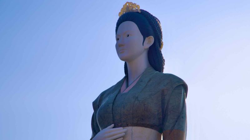 A close-up view of the Lady Suro sculpture in red robes, crowned, gazing upwards in Lady Suro Memorial Park.