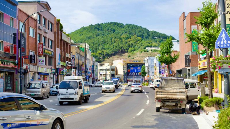 The main road cuts through Mukho Port, bordered by three-story stores on either side, with a tree-covered hillside looming in the distance.