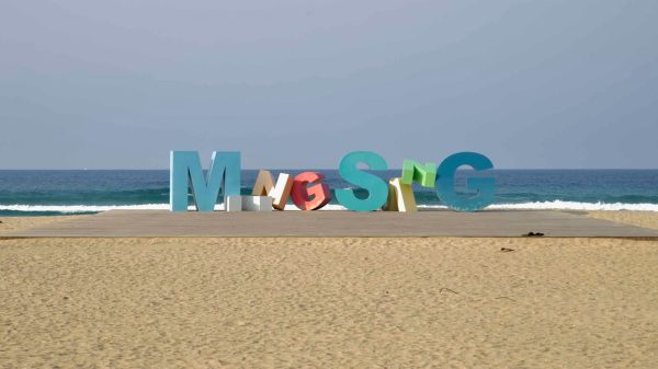 A playful sculpture spelling “Mangsang” stretches across a beach platform, set against low waves rolling onto the golden sand of Mangsang Beach.