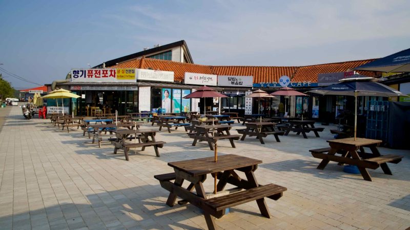 Rows of wooden picnic tables and umbrellas stand ready outside a cluster of restaurants and cafes near Mangsang Beach, inviting visitors to dine in the sun.