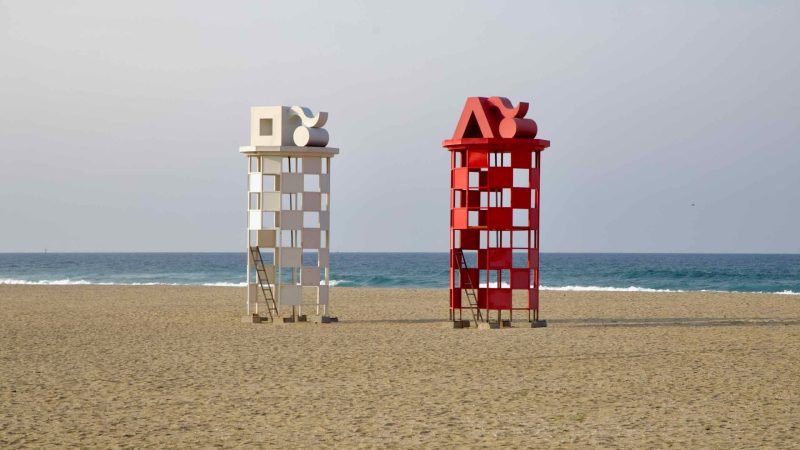 Two tower-like sculptures in red and white checkerboard patterns stand side by side on the sands of Mangsang Beach, facing calm waves of the East Sea.