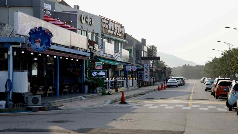 A row of seafood restaurants and local shops lines a quiet street near Mangsang Beach, with parked cars, pine trees, and hazy mountains in the background.