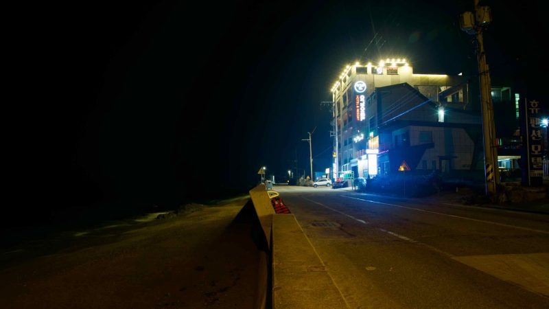 A brightly lit motel along the coastal road near Ganggu Port illuminates the night, with a dark beach and the East Sea stretching out below.