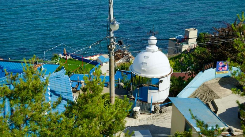 Nongoldam Street, featuring vibrant blue rooftops and an old lighthouse structure, with the serene East Sea in the background.