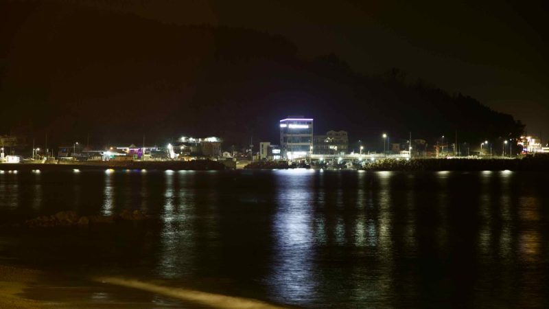 Nighttime view of restaurants on the outskirts of Ganggu Port, with bright lights along the coastal roadway, the dark East Sea and waves in the foreground under a black sky.