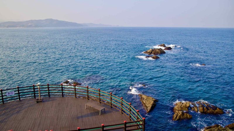 Overlooking the East Sea from a rock observatory, the semicircle platform of the Chogok Yonggul Cave Hiking Trail captures rocks above waves and distant mountains.