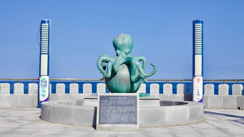 A whimsical octopus sculpture, perched atop a ball near the East Sea at Mukho Port, stands out against the backdrop of a clear blue sky, adding a playful touch to the coastal landscape.