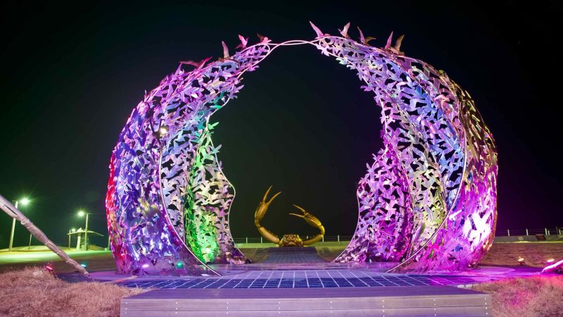 In Haeparang Park, colorful lights illuminate a metal sculpture of interconnected birds in flight, with a large snow crab sculpture in the background against the black night sky.