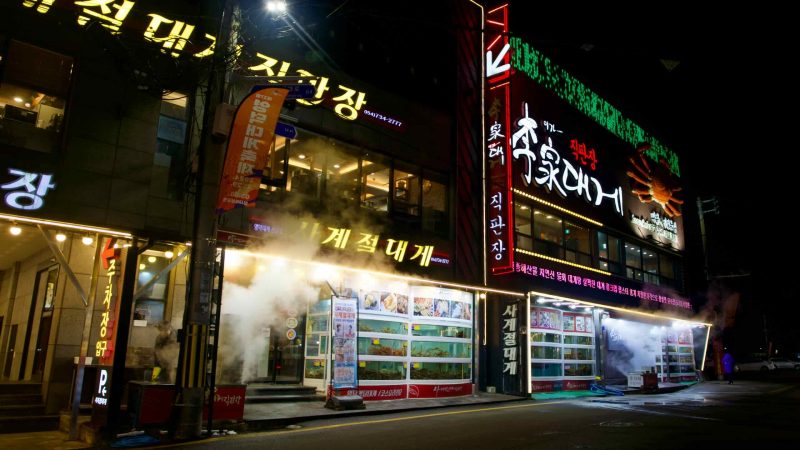 Outside a three-story snow crab restaurant in Ganggu Port, steam from snow crab cookers fills the air, with a snow crab statue emblazoned on the side and tanks in front.
