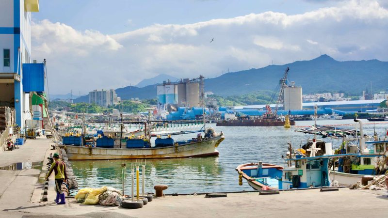 A squid fishing boat adorned with large lightbulbs for night fishing rests near the bustling fishing market, with a worker strolling along the port, embodying the daily rhythm of maritime life.