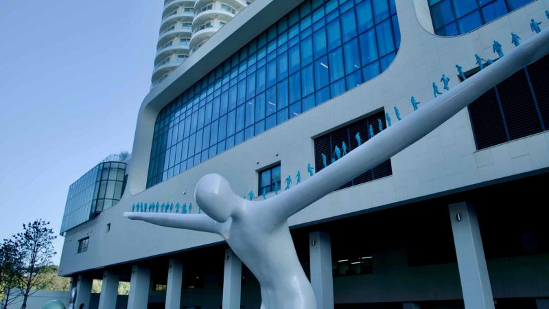 A contemporary sculpture with outstretched arms stands in front of the Cassia Sokcho Hotel, blending modern art with the hotel's sleek architectural design at Daepo Port.