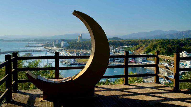 A crescent moon sculpture frames a panoramic view of Daepo Port from the observatory, offering a unique vantage of the harbor, coastline, and surrounding mountains.