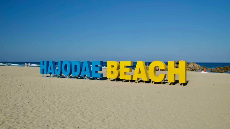 The vibrant Hajodae Beach sign glows under the afternoon sun on the golden sands of Yangyang County's coastline, offering a serene backdrop of the East Sea.