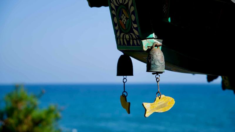 A close-up of a temple bell with a fish-shaped wind chime hanging from the roof of Hongnyeonam Hermitage, with the East Sea as a tranquil backdrop.