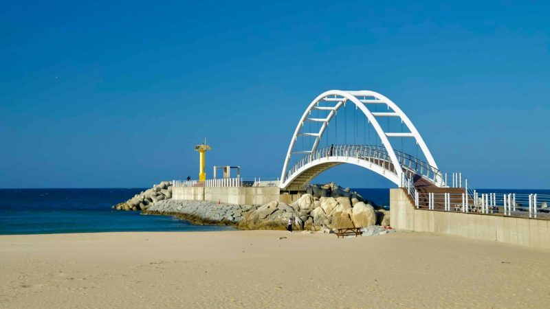 A scenic view of the arched bridge leading to the yellow lighthouse at Namae Sunrise Observatory, surrounded by sandy beaches and rocky shorelines.