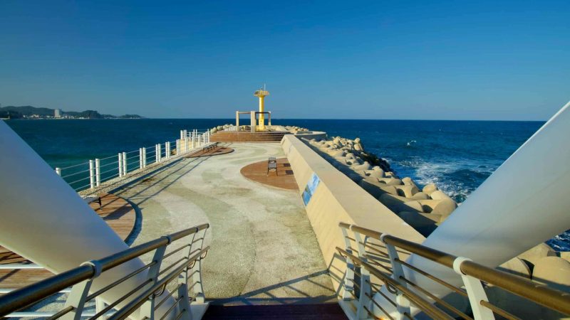 The landing area of Namae Sunrise Observatory features a paved walkway, a yellow lighthouse, and panoramic views of the East Sea under a clear sky.