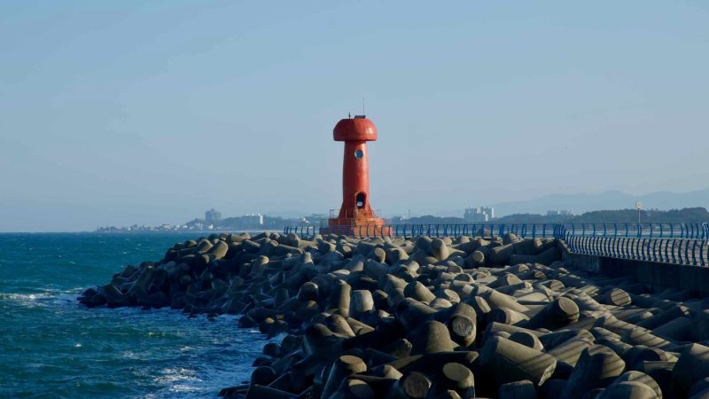 A red lighthouse stands atop a breakwater at Namae Port, surrounded by tetrapods and offering a striking contrast against the deep blue East Sea.