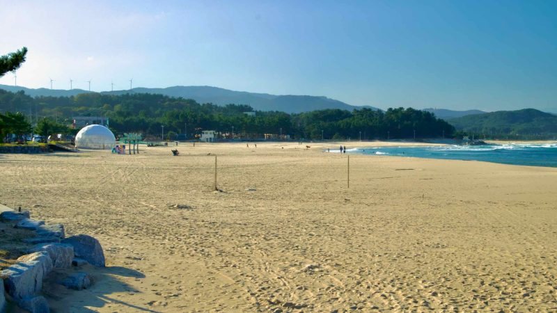 A tranquil scene of Namae Beach featuring expansive sandy shores, gentle waves, and a backdrop of forested hills under a clear sky.