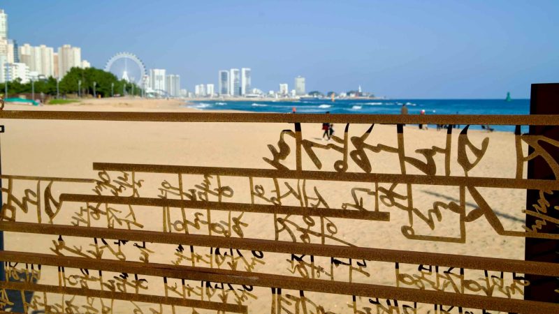 A decorative metal gate near the entrance of the Oeongchi Sea Fragrance Trail frames a scenic view of Sokcho Beach, with distant city buildings and the Ferris wheel.