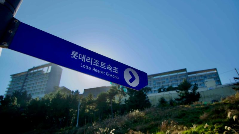 A directional sign pointing to Lotte Resort Sokcho stands against a clear sky, with the resort building visible in the background atop a coastal hill.