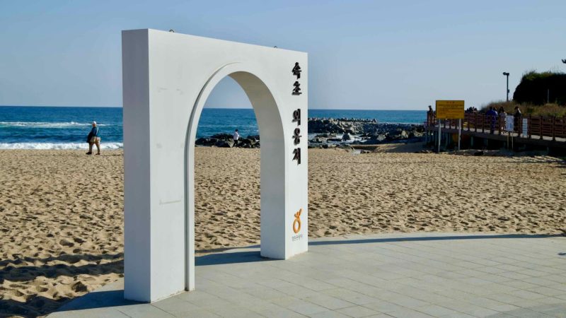 The white archway marks the entrance to Oeongchi Beach, a serene stretch of sand and surf along the East Sea, with nearby boardwalks and coastal views.