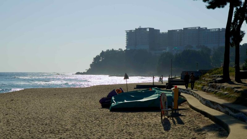 A serene view of Sokcho Beach during sunset, with waves shimmering in the light and the Lotte Resort perched on a forested hill in the background.