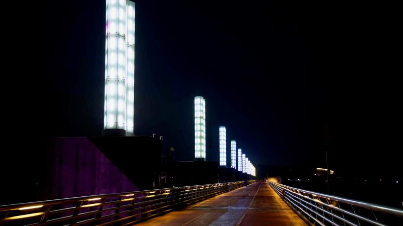 A nighttime view down the top deck of Yeoju Weir shows a line of illuminated pillars modeled after a water clock, glowing over the bike and pedestrian path.