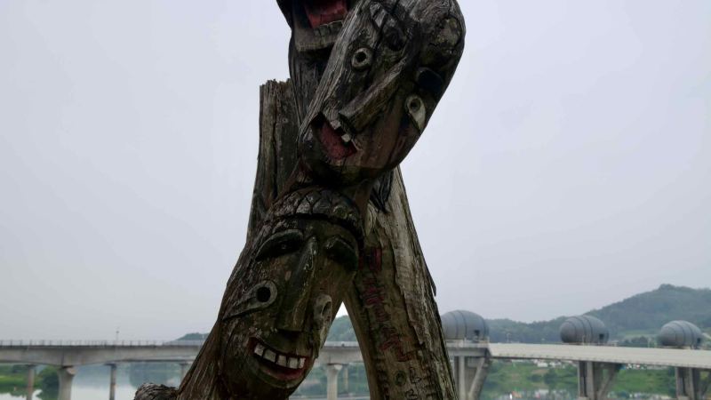 Expressive carved wooden totems overlook the South Han River, with Ipo Weir’s egg-shaped gatehouses and cascading spillway visible in the distance.