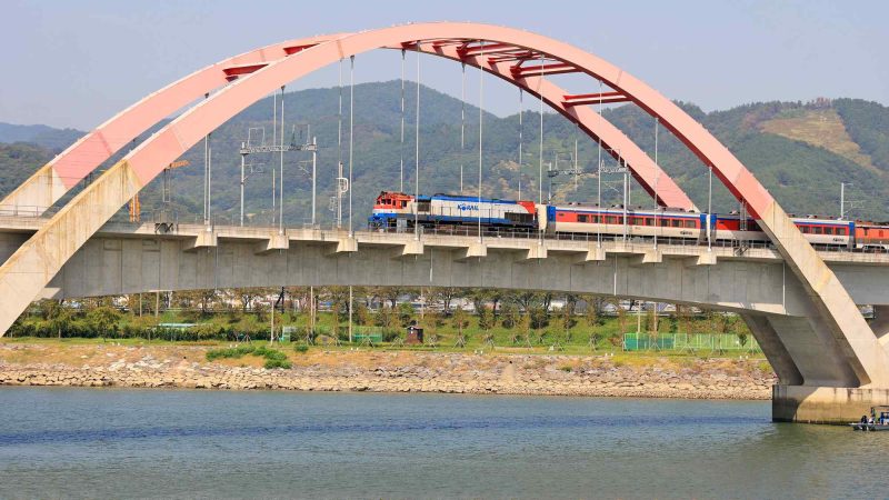 A KORAIL train passes over a modern arched railroad bridge spanning the Seomjin River.