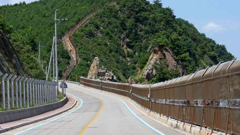 A coastal road near Geojin Port winds 53 meters up through a mountain pass, flanked by a military fence and the rocky coast of the East Sea.