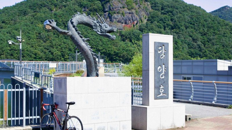 Road bike leans against a dragon statue at Damyang Lake, backed by forested hills.
